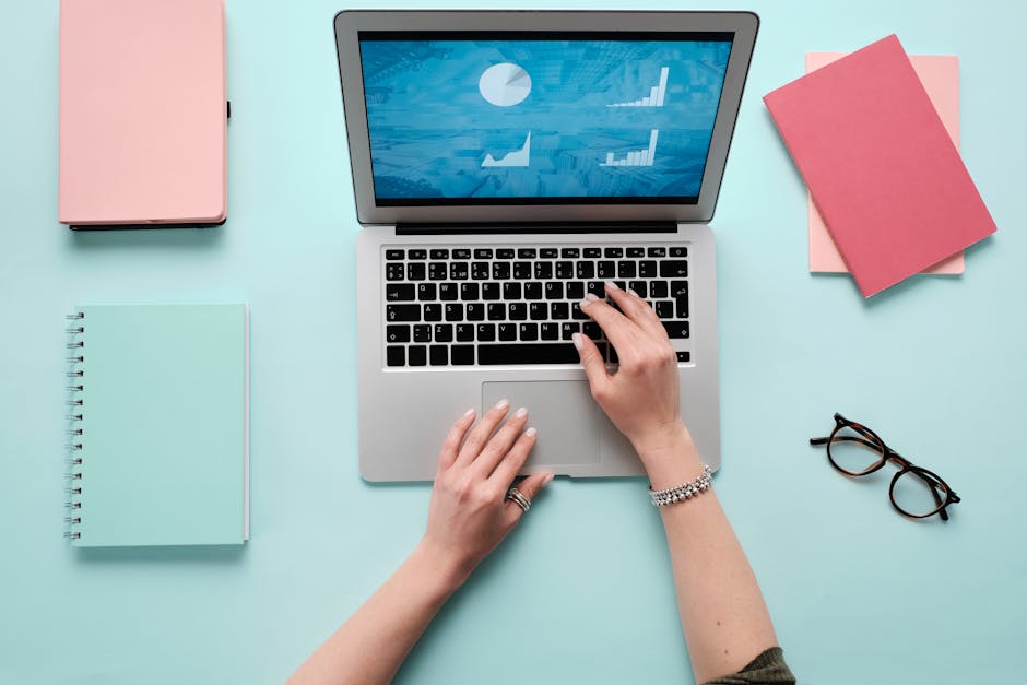 Hands typing on a laptop displaying data charts, with pink and teal office accessories.